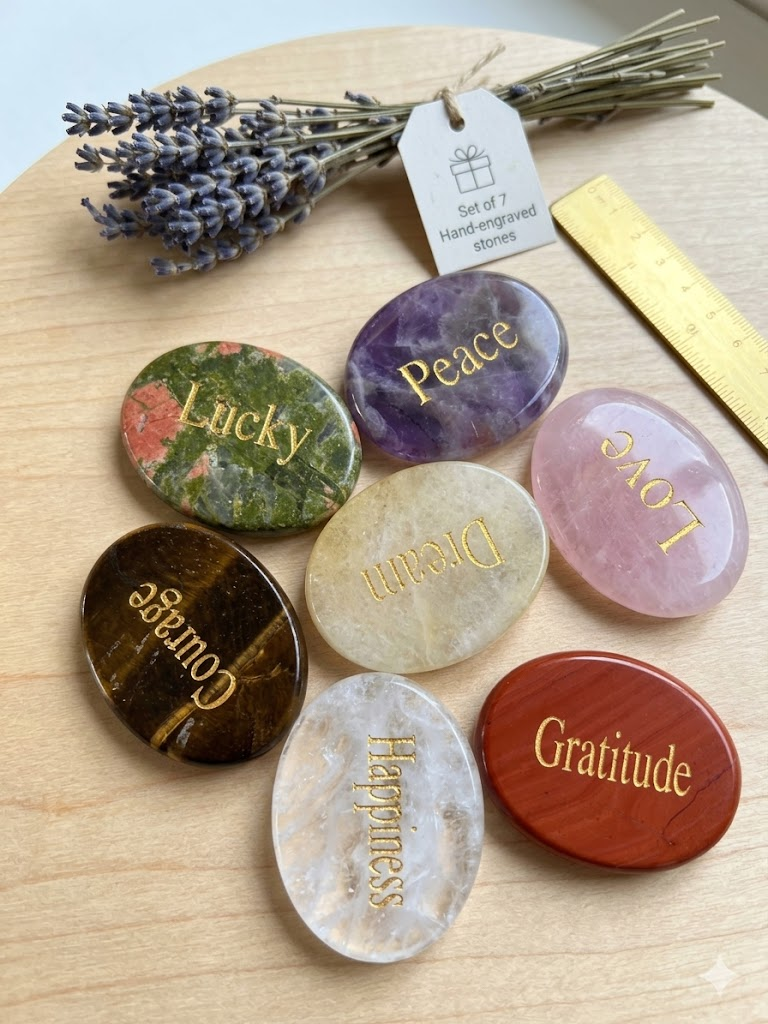 A premium collection of 7 natural engraved crystal worry stones featuring gold lettering (Peace, Love, Lucky, Courage, Happiness, Dream, Gratitude) arranged elegantly on a clean, minimalist outdoor setting under warm, natural sunlight.