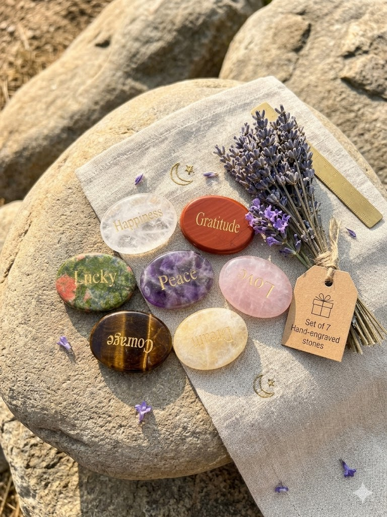 A premium collection of 7 natural engraved crystal worry stones featuring gold lettering (Peace, Love, Lucky, Courage, Happiness, Dream, Gratitude) arranged elegantly on a clean, minimalist outdoor setting under warm, natural sunlight.