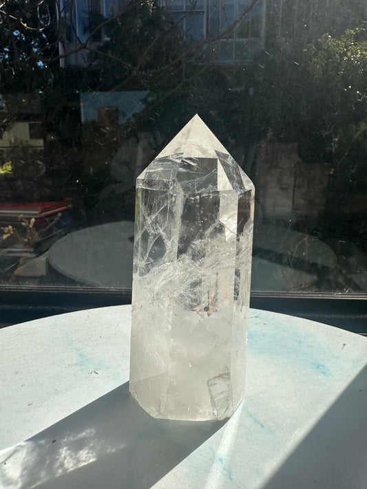 Brazilian clear quartz tower in sunlight, with a polished pointed top, transparent upper section, white internal veils and a milky base. Exact 634g piece, approximately 14cm tall.
