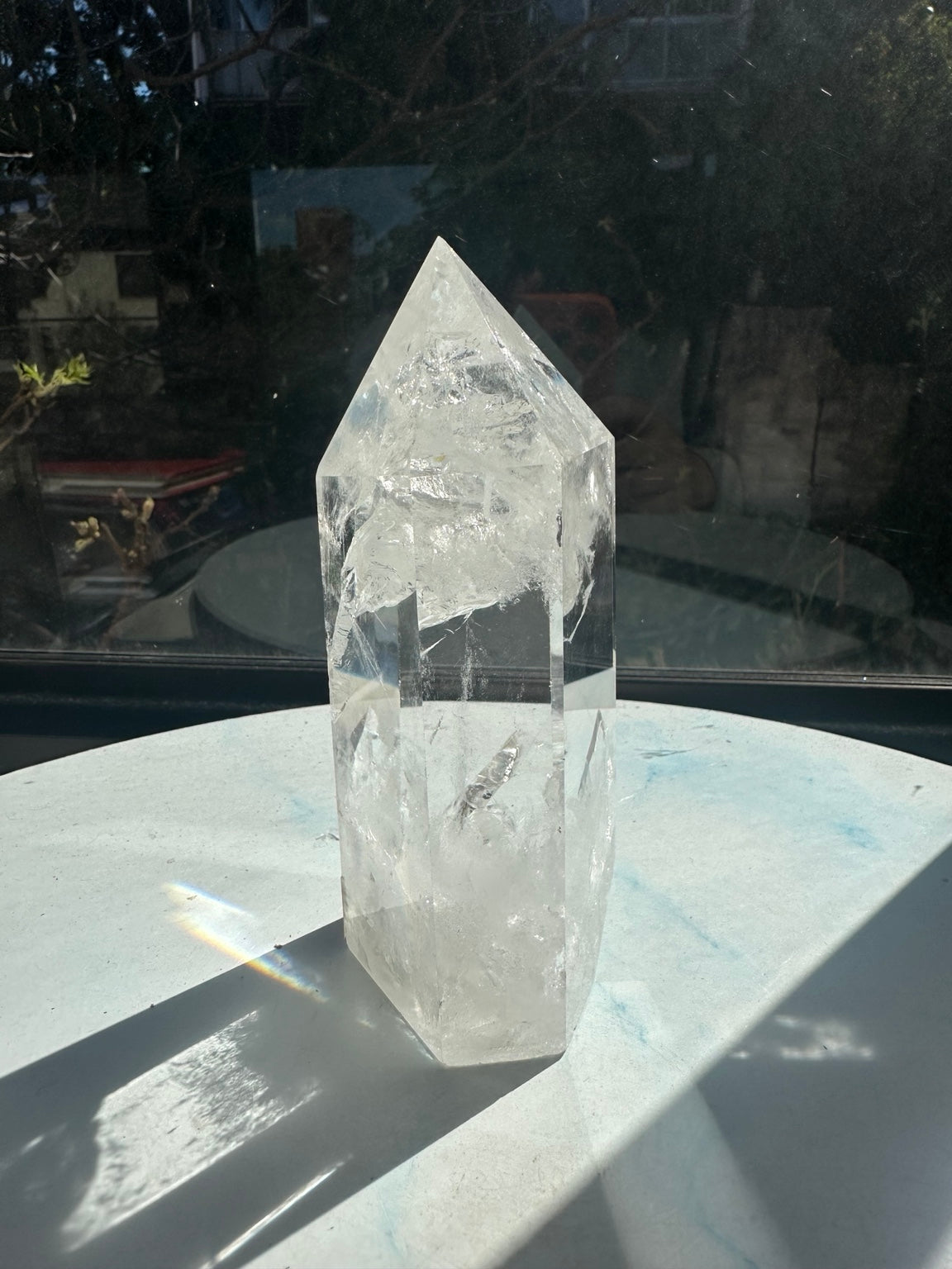 Side view of quartz tower #2 showing the full pointed top, internal fracture textures and a rainbow light patch cast onto the table beside the crystal.