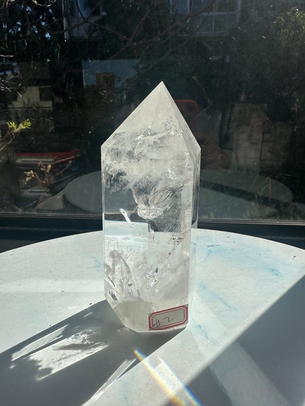 Front angled view of the same clear quartz tower with a red #2 label at its base and rainbow light visible on the surrounding surface in strong sunlight.
