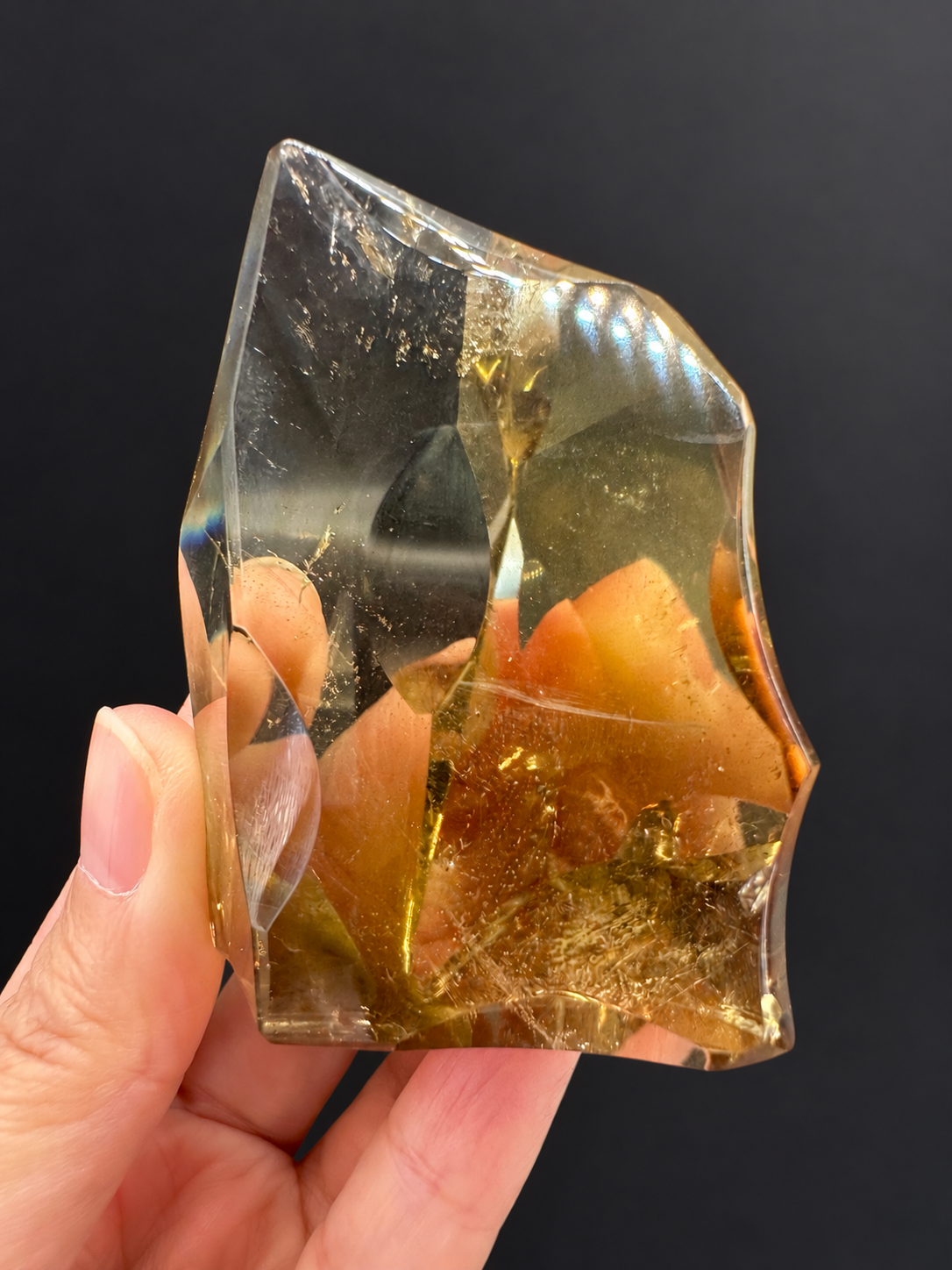 Brazilian citrine freeform No.7, 117g, approx. 7 × 5cm; rotated front view showing golden edges, fine internal veils and the curved upper edge, held in the original hand against a charcoal background.