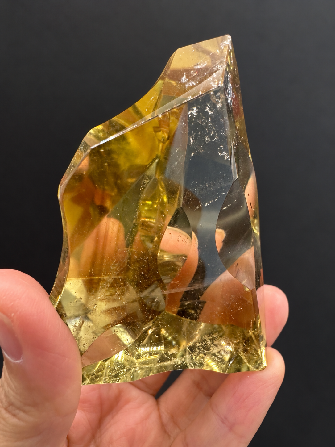 Brazilian citrine freeform No.7, 117g, approx. 7 × 5cm; angled reverse view showing the golden base and intersecting polished curved facets, held in the original hand against a charcoal background.