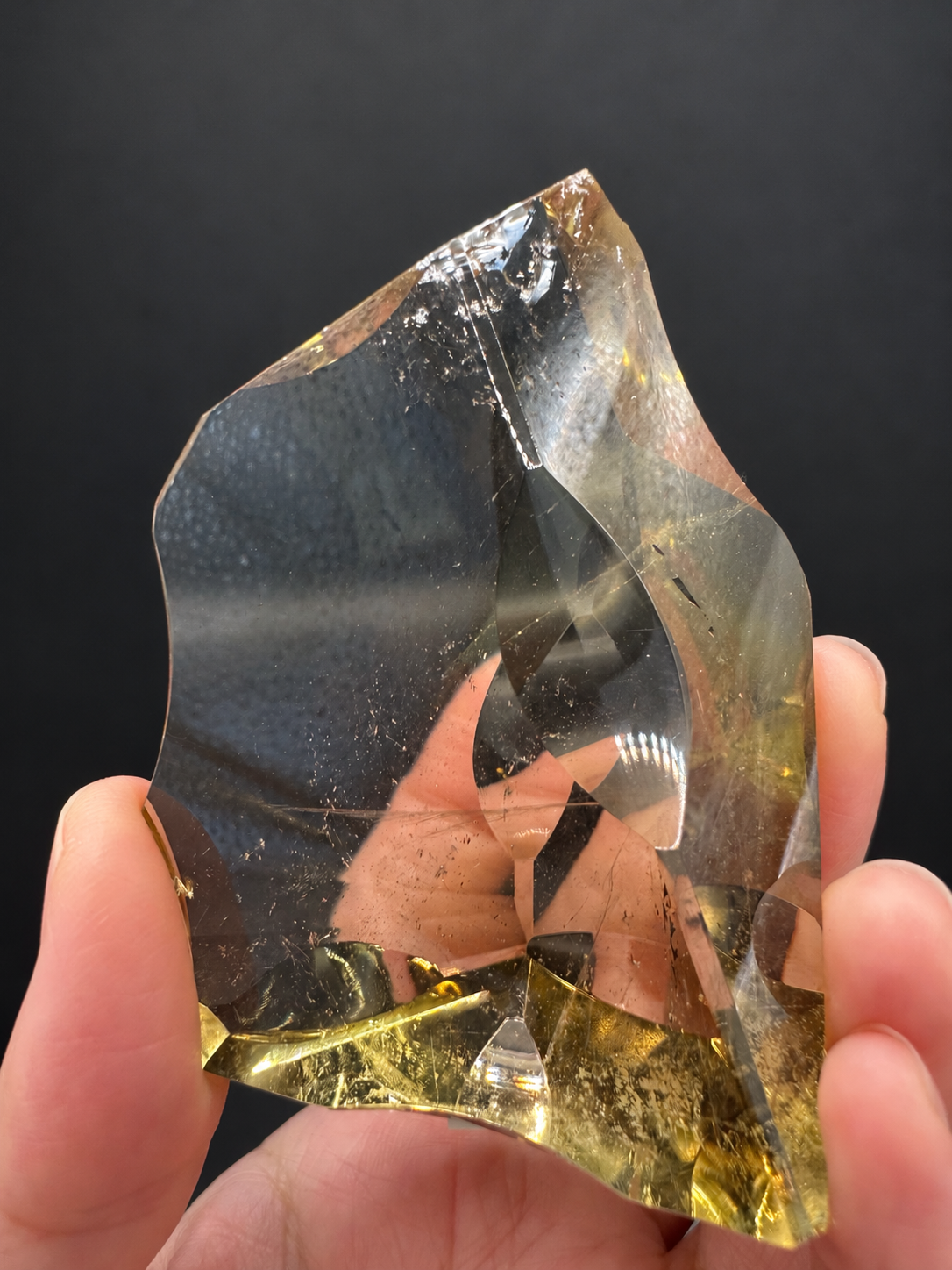 Brazilian citrine freeform No.7, 117g, approx. 7 × 5cm; reverse front view showing a broad transparent window, shadow contrast and fine internal specks, held in the original hand against a charcoal background.