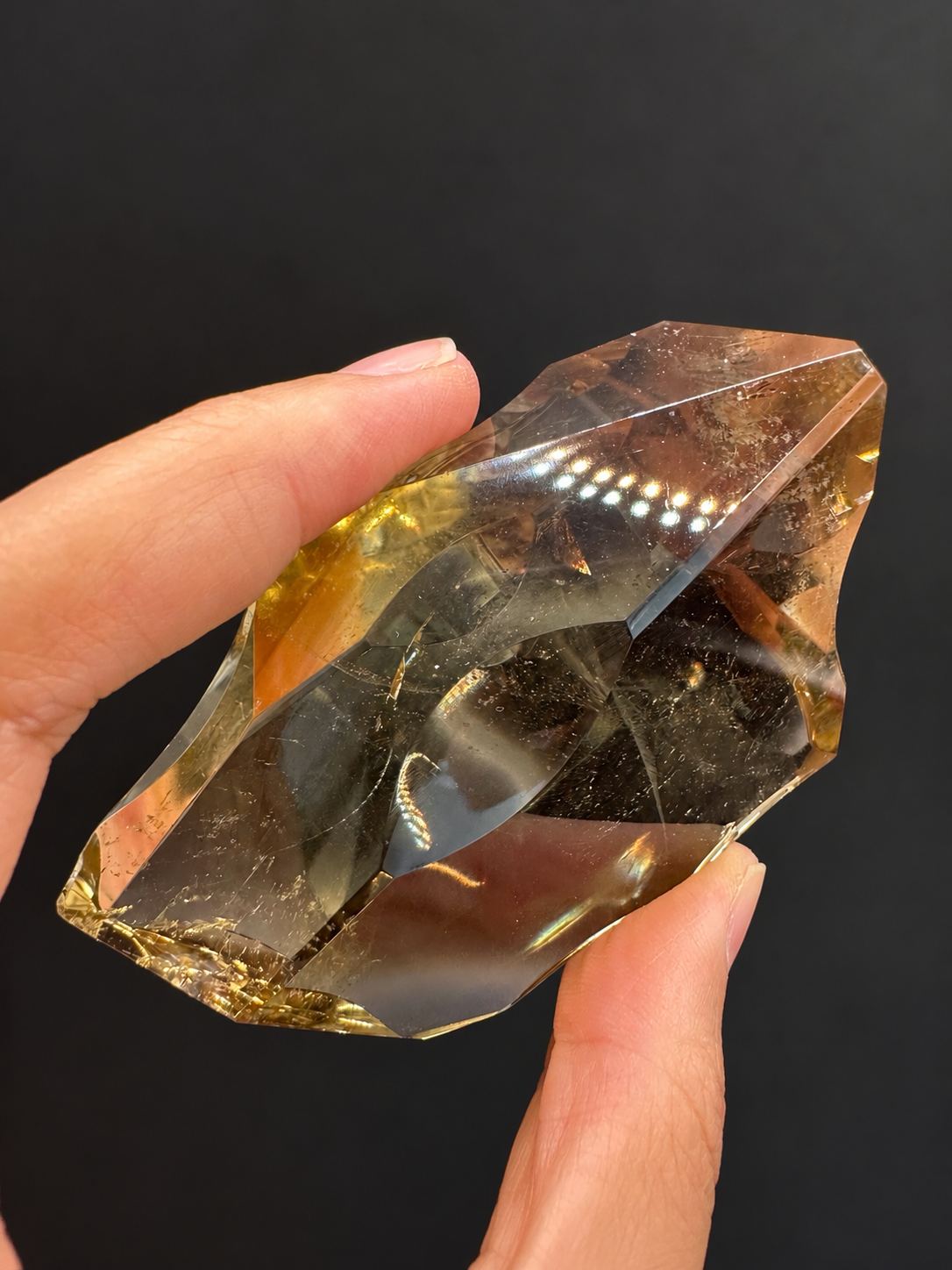 Brazilian citrine freeform No.7, 117g, approx. 7 × 5cm; tilted view showing curved facets, shadow contrast and original reflected lights, held in the original hand against a charcoal background.
