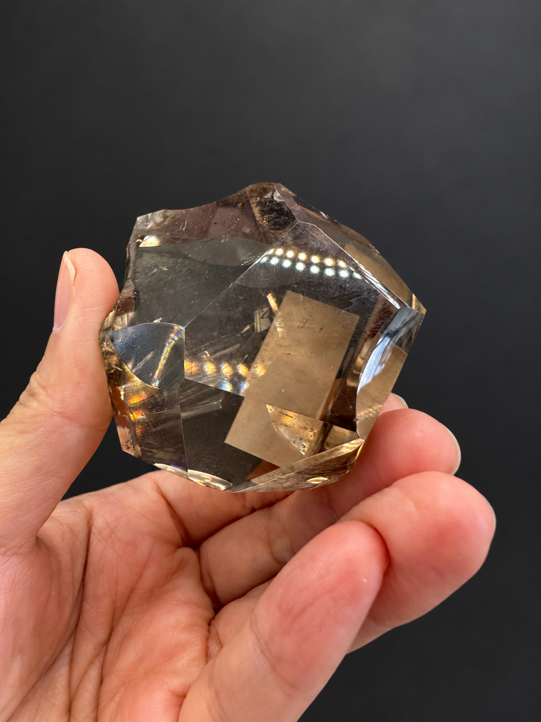 Brazilian citrine freeform No.8, 134g, approx. 5 × 5cm; rotated palm-held view of intersecting polished facets and champagne tones, held in the original hand against a charcoal background.