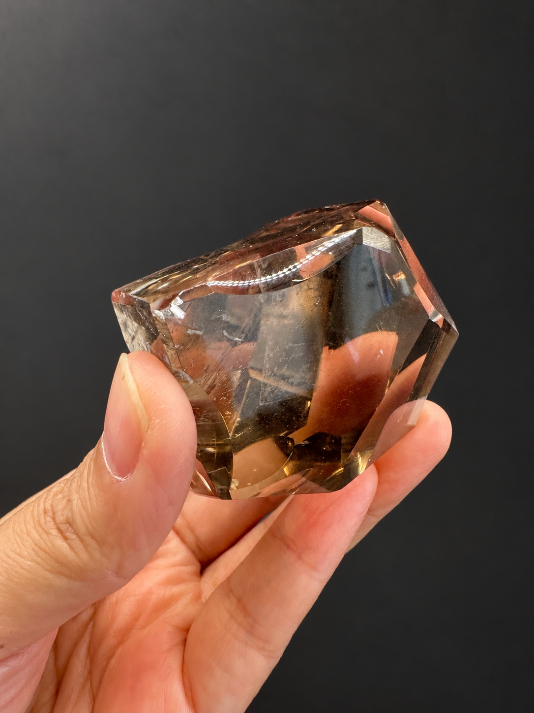 Brazilian citrine freeform No.8, 134g, approx. 5 × 5cm; angled view showing the curved upper edge and fingers visible through the crystal, held in the original hand against a charcoal background.