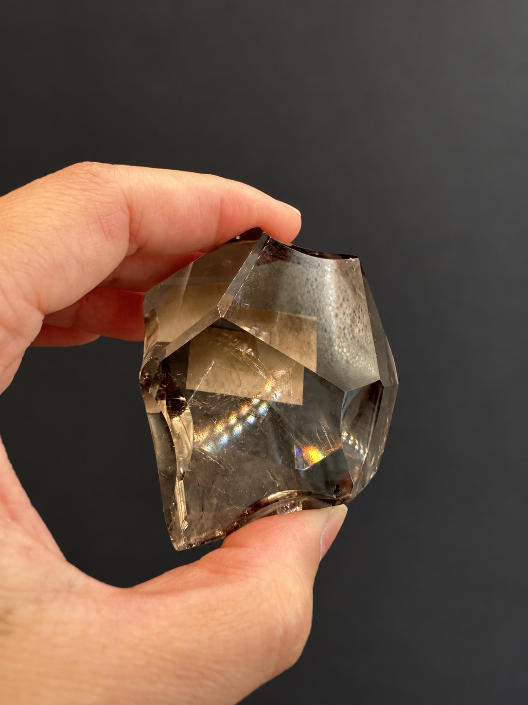 Brazilian citrine freeform No.8, 134g, approx. 5 × 5cm; pinch-held view showing curved side facets and original reflected light, held in the original hand against a charcoal background.