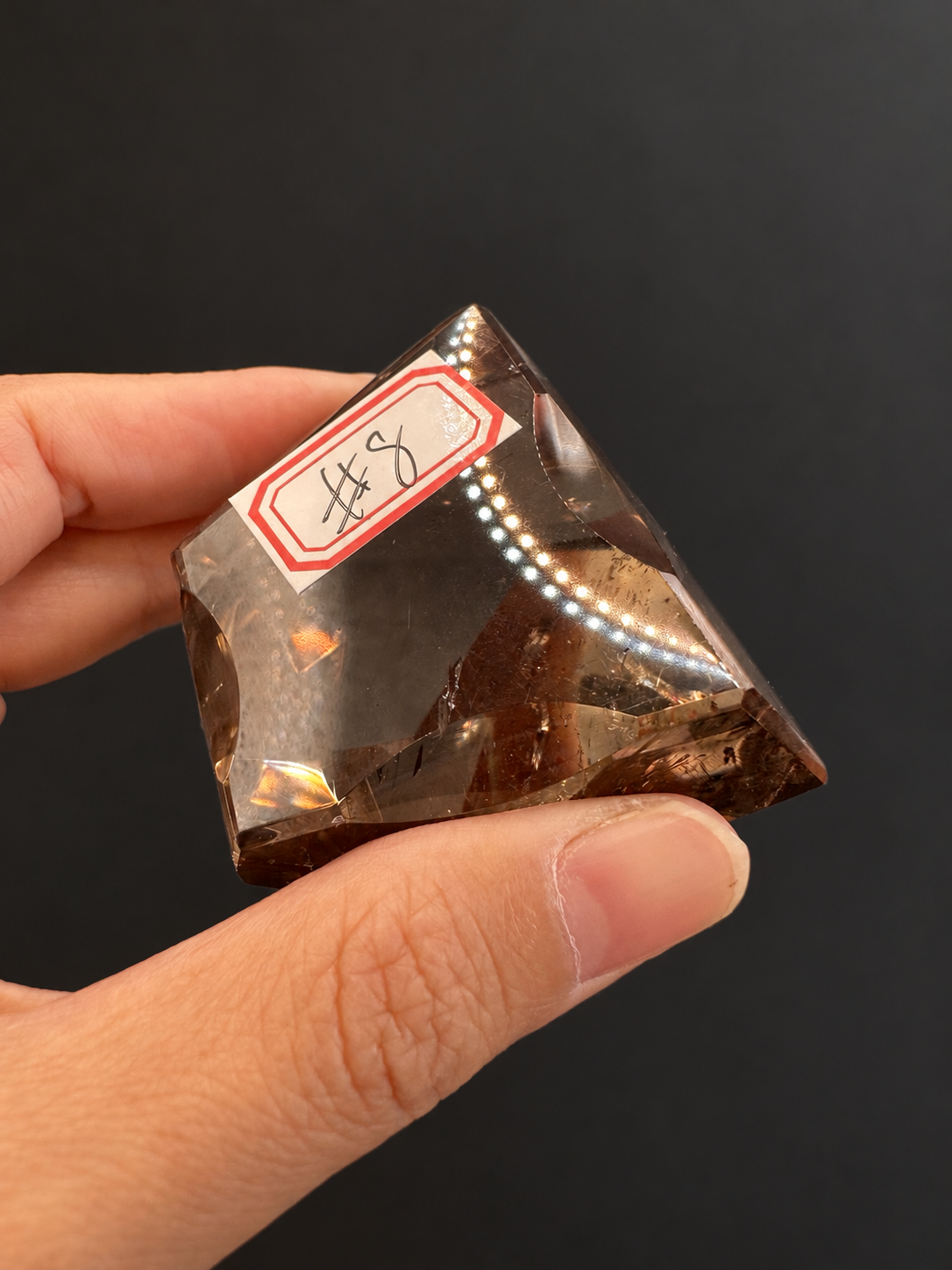 Brazilian citrine freeform No.8, 134g, approx. 5 × 5cm; tilted labelled No.8 view showing a broad transparent face and original reflections, held in the original hand against a charcoal background.