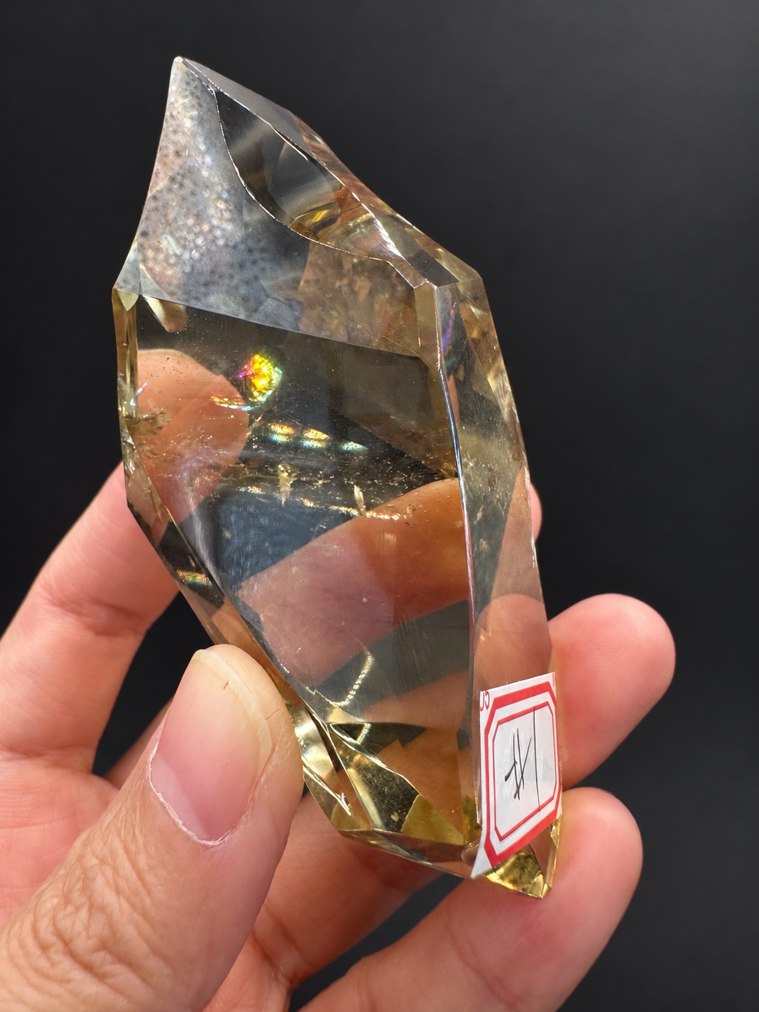 Brazilian citrine freeform No.1, 100g, approximately 8 × 4.4cm; rotated broad face with a brighter rainbow patch and original side label, held in the original hand against a charcoal background.