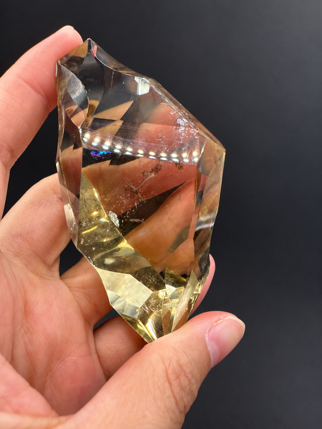 Brazilian citrine freeform No.1, 100g, approximately 8 × 4.4cm; reverse broad face with golden lower facets, wispy fractures and original dotted light reflections, held in the original hand against a charcoal background.