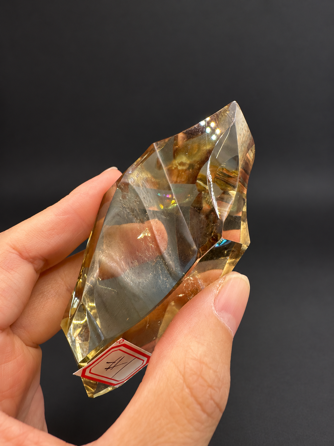 Brazilian citrine freeform No.1, 100g, approximately 8 × 4.4cm; angled face with sweeping concave cuts, original label and small coloured flashes, held in the original hand against a charcoal background.