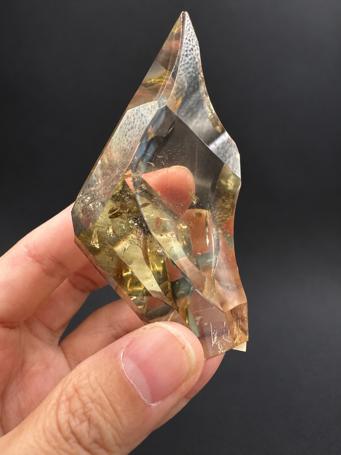 Brazilian citrine freeform No.1, 100g, approximately 8 × 4.4cm; opposite side showing the pointed tip, concave curved facets and original white textured reflections, held in the original hand against a charcoal background.