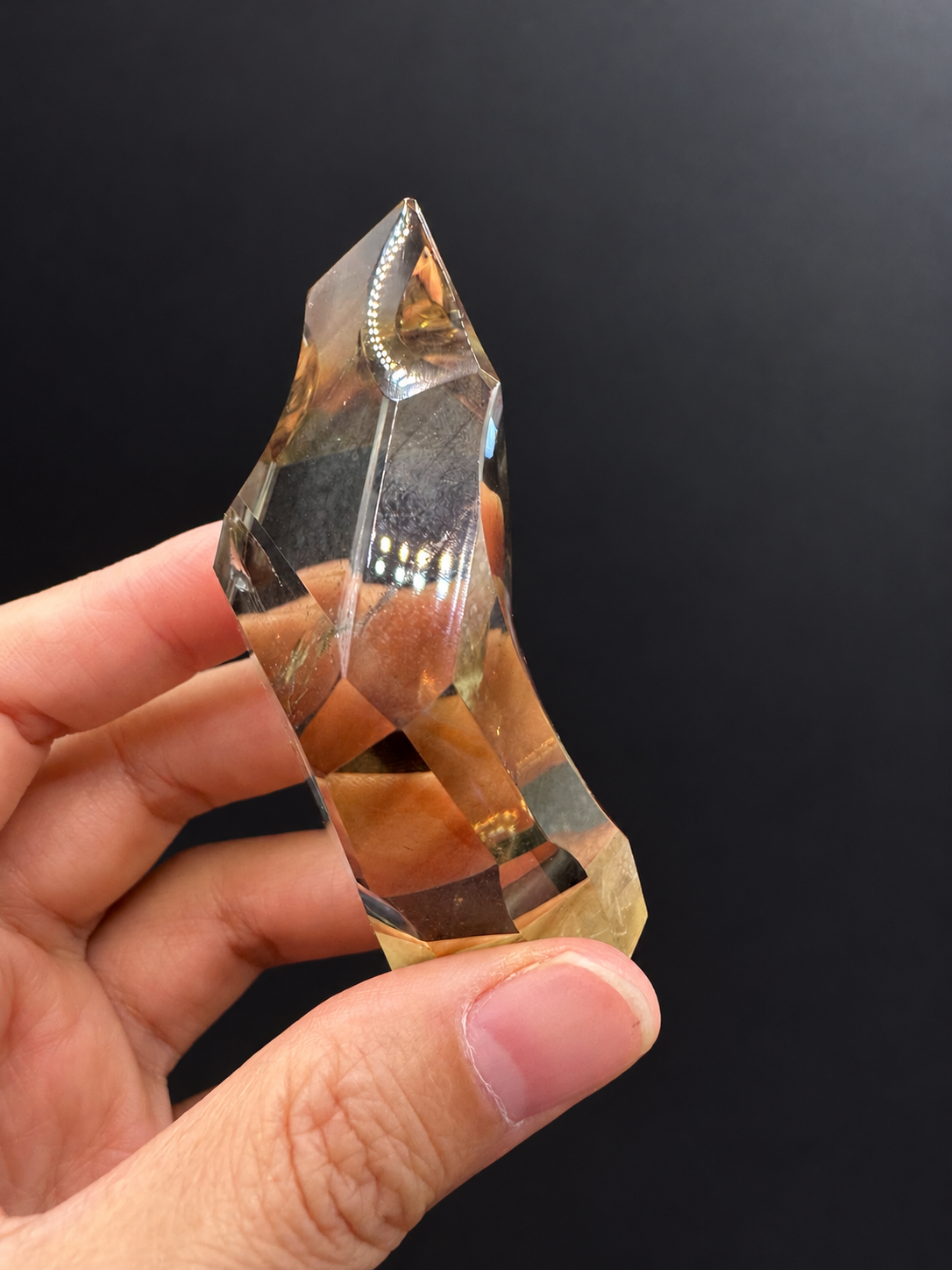 Brazilian citrine freeform No.1, 100g, approximately 8 × 4.4cm; slim edge-on profile with clear curved cuts and golden lower facets, held in the original hand against a charcoal background.
