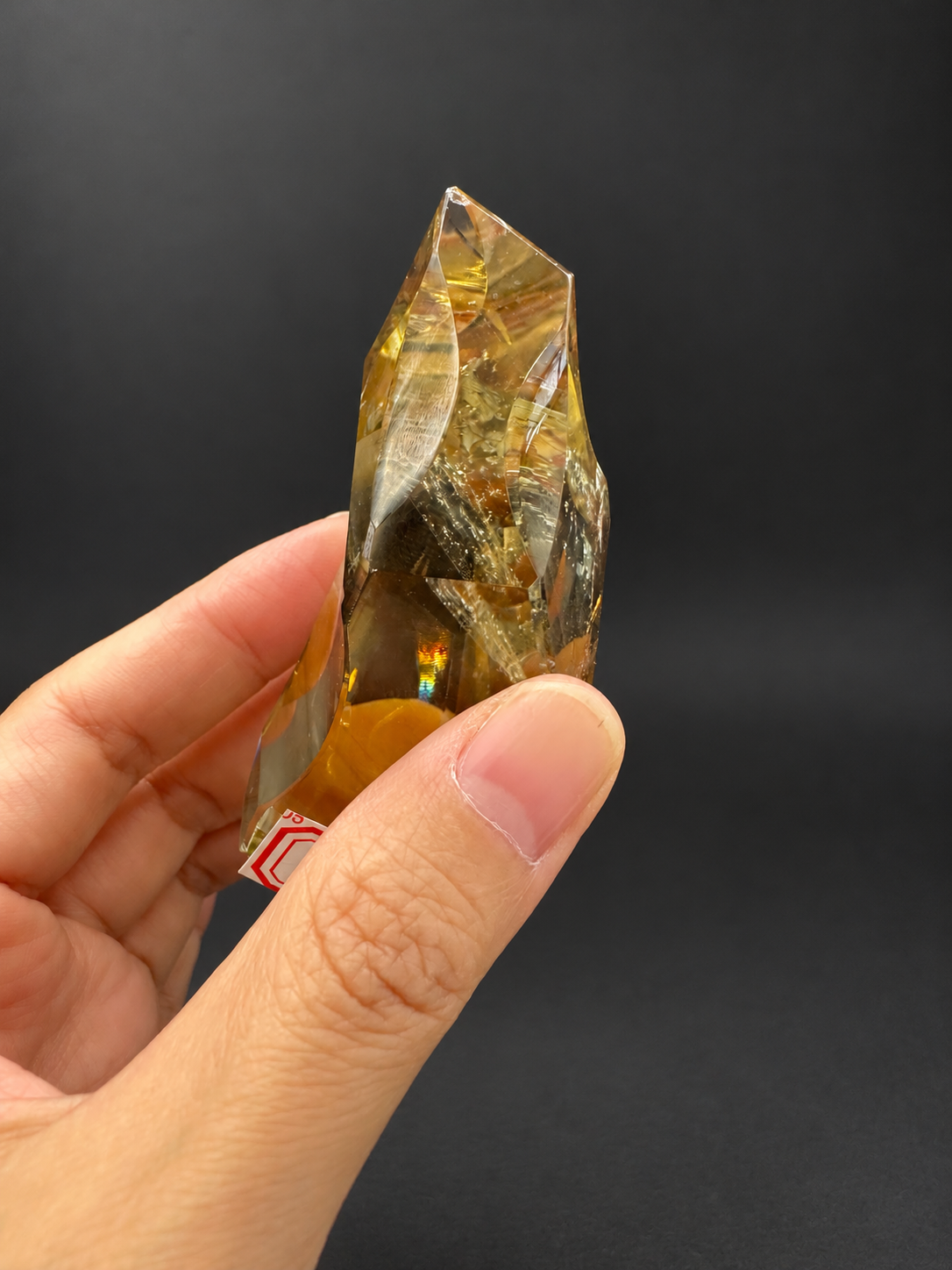 Brazilian citrine freeform No.1, 100g, approximately 8 × 4.4cm; thin oblique side showing golden interior, visible fractures and a small rainbow reflection, held in the original hand against a charcoal background.