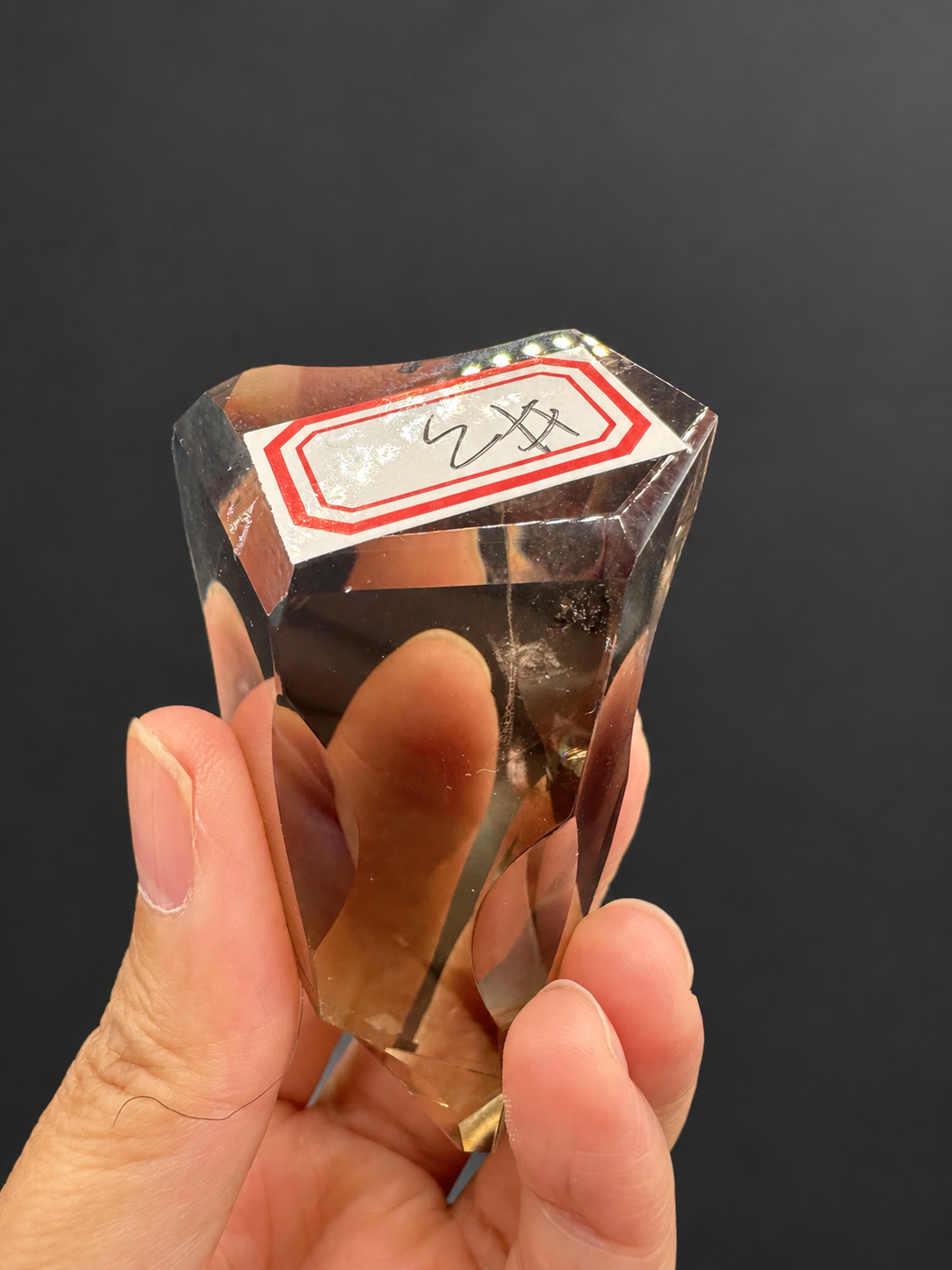 Brazilian citrine freeform No.3, 89g, approximately 7 × 4cm; labelled identification view with the original red-bordered sticker and visible clear window, held in the original hand, against a charcoal background.