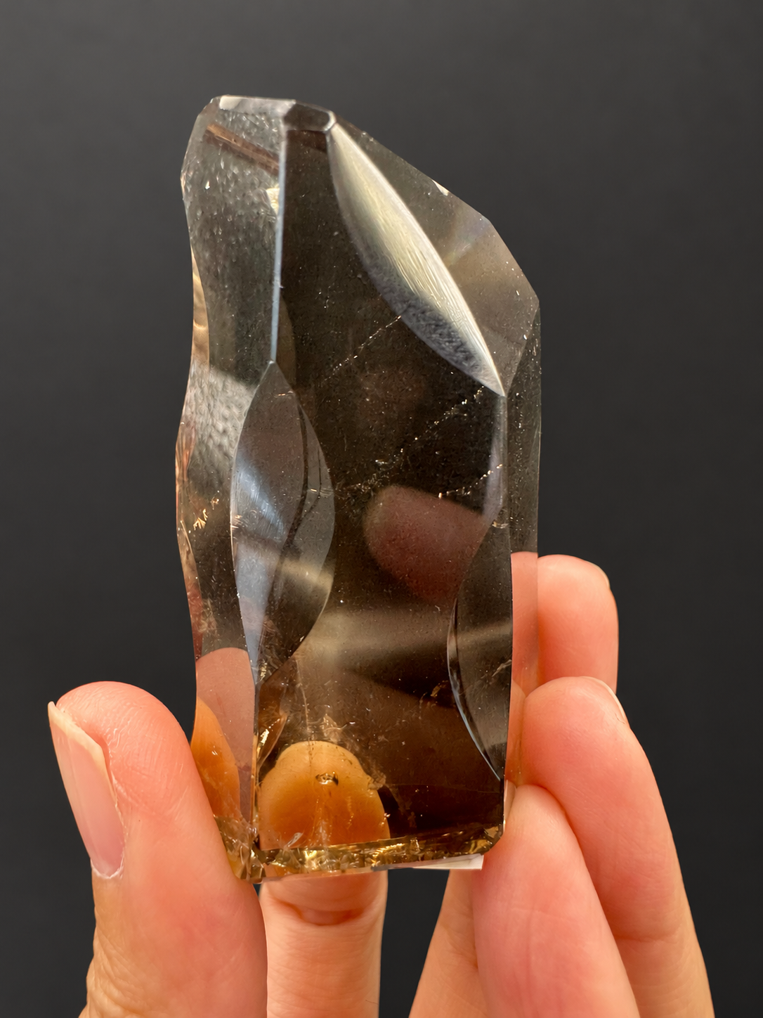 Brazilian citrine freeform No.3, 89g, approximately 7 × 4cm; narrower oblique view showing sculpted side cuts, transparent body and golden base, held in the original hand, against a charcoal background.