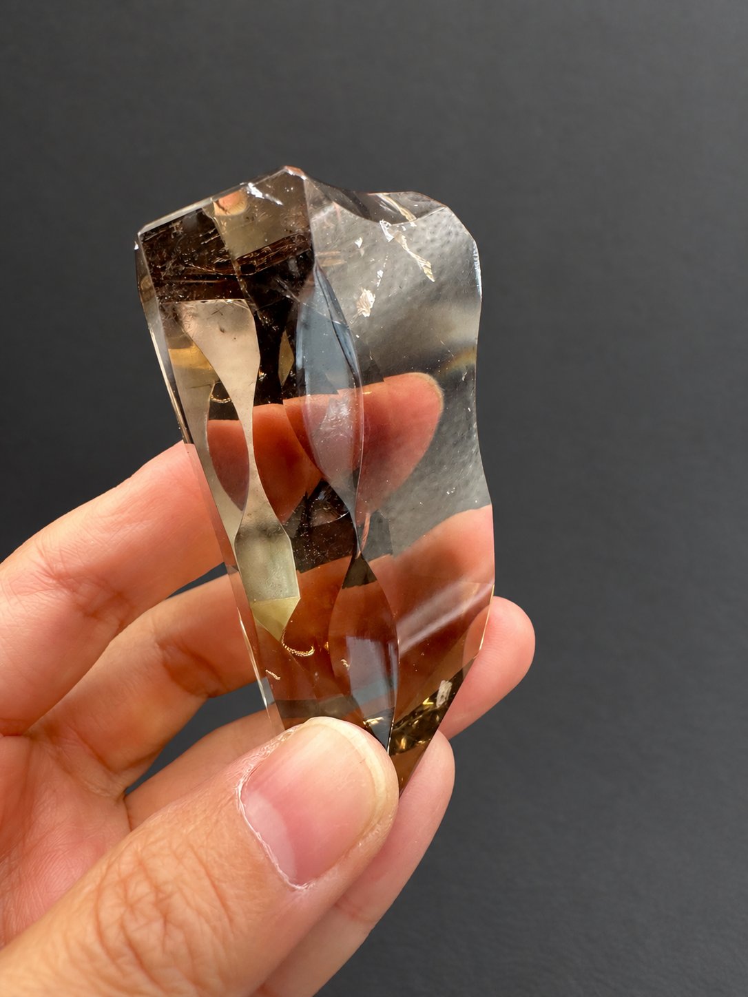 Brazilian citrine freeform No.3, 89g, approximately 7 × 4cm; reverse angled view showing flowing concave cutwork and original textured light reflections, held in the original hand, against a charcoal background.