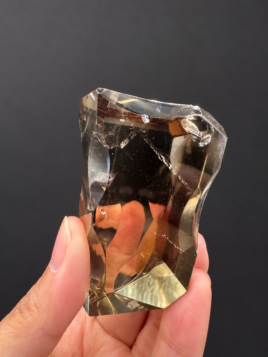 Brazilian citrine freeform No.3, 89g, approximately 7 × 4cm; broad front view showing an irregular upper rim, visible diagonal fractures and angular gold lower facets, held in the original hand, against a charcoal background.