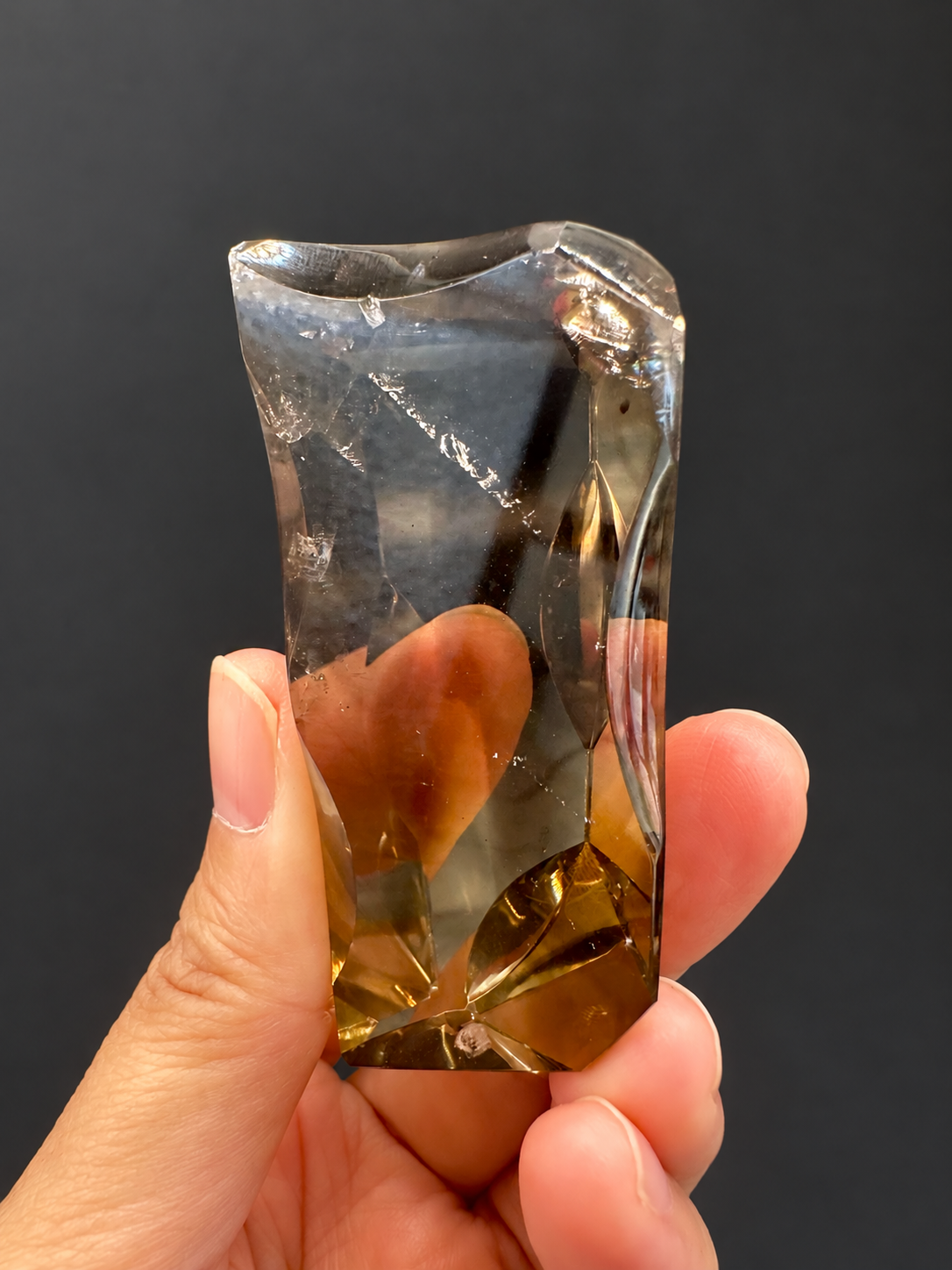 Brazilian citrine freeform No.3, 89g, approximately 7 × 4cm; slightly rotated front face showing transparency, small specks and the curved side profile, held in the original hand, against a charcoal background.