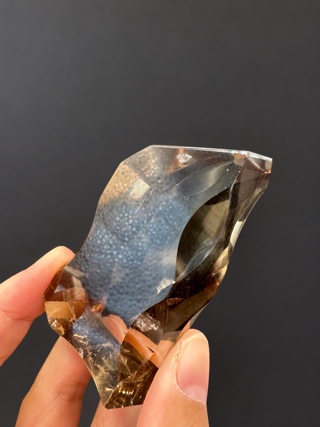 Brazilian citrine freeform No.3, 89g, approximately 7 × 4cm; tilted side view with large concave cuts and original dotted lamp reflections, held in the original hand, against a charcoal background.