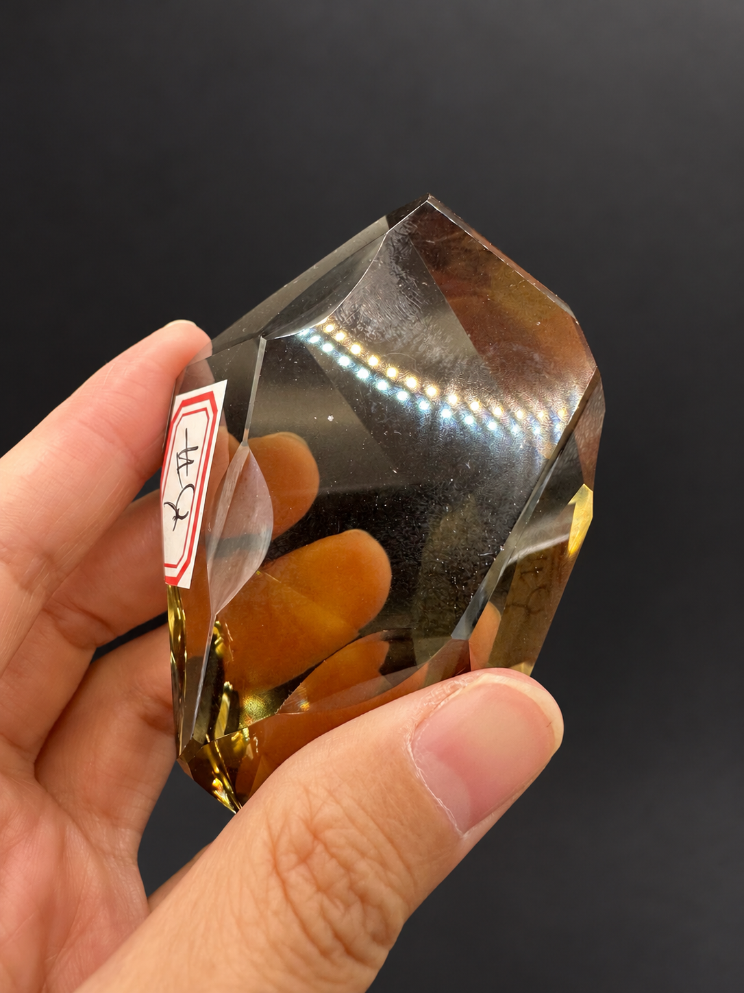 Brazilian citrine freeform No.4, 116g, approximately 8 × 5cm; labelled rotated face with original dotted light reflections, held in the original hand against a charcoal background.