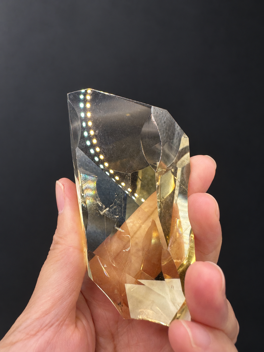 Brazilian citrine freeform No.5, 111g, approximately 9 × 4.2cm; broad clear front face with sweeping curved top, thin outline and original dotted light reflections, held in the original hand against a charcoal background.