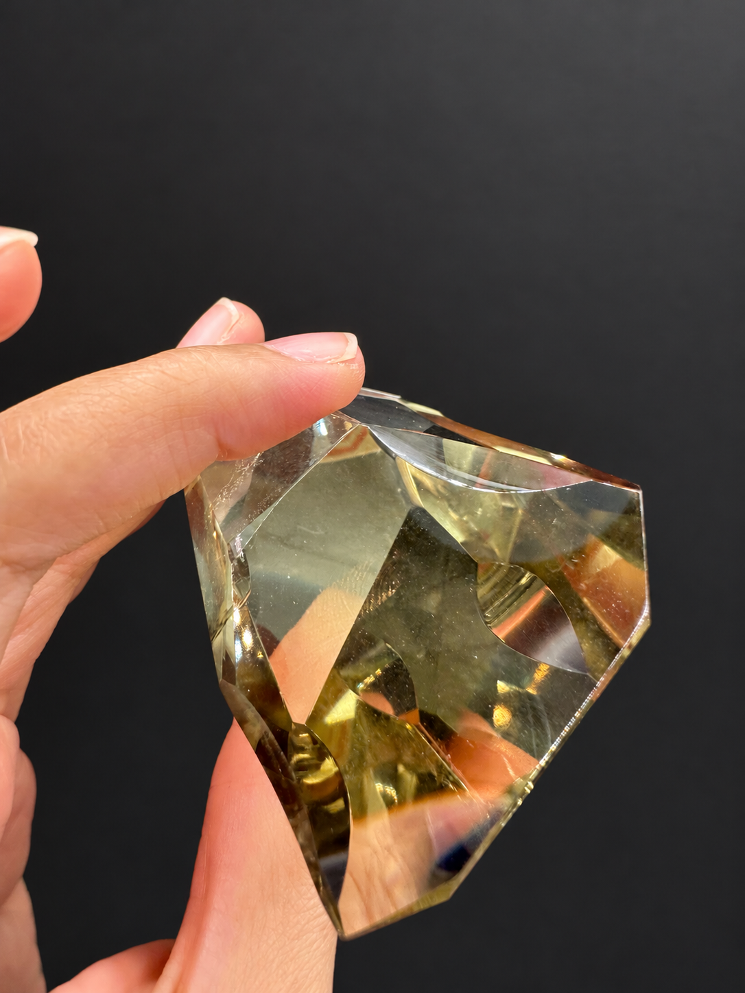 Brazilian citrine freeform No.5, 111g, approximately 9 × 4.2cm; tilted upper and depth view held between original fingers, showing curved and planar facets, held in the original hand against a charcoal background.