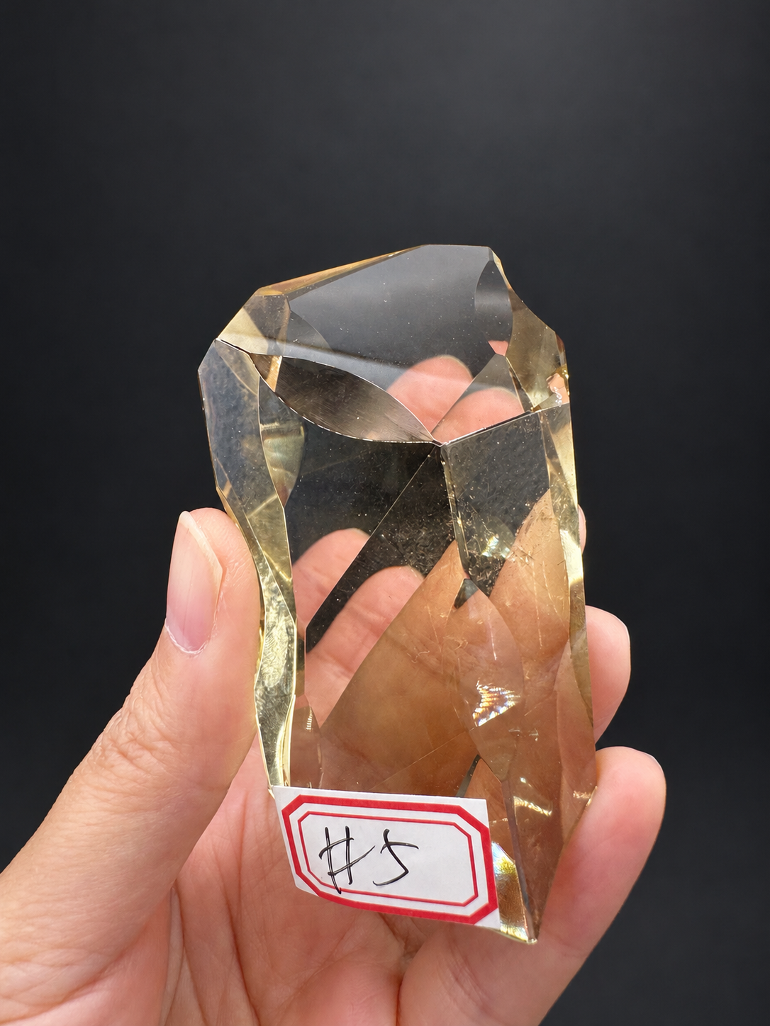 Brazilian citrine freeform No.5, 111g, approximately 9 × 4.2cm; labelled No.5 face showing transparent champagne windows and intersecting facets, held in the original hand against a charcoal background.