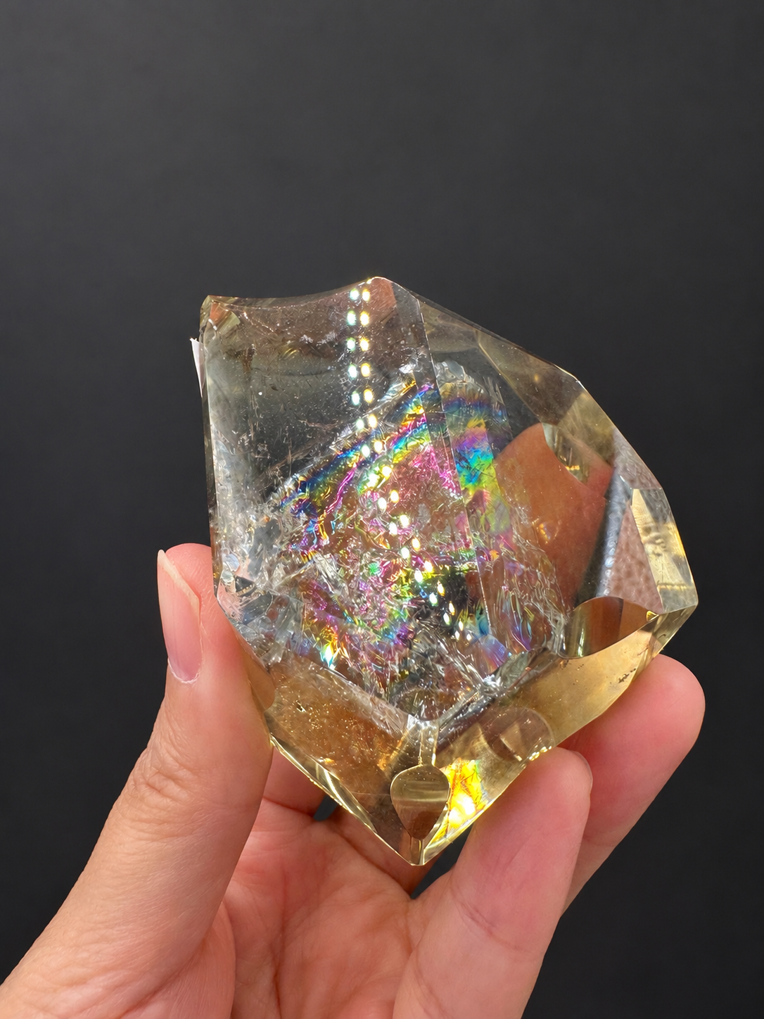Brazilian citrine freeform No.6, 203g, approximately 6.3 × 5.8cm; broad front face with multicoloured internal rainbows and the sculpted concave top, held in the original hand against a charcoal background.