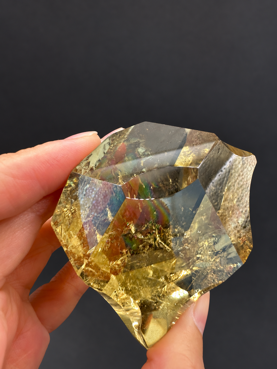 Brazilian citrine freeform No.6, 203g, approximately 6.3 × 5.8cm; tilted broad curved facets with golden colour and subtle central rainbow flashes, held in the original hand against a charcoal background.