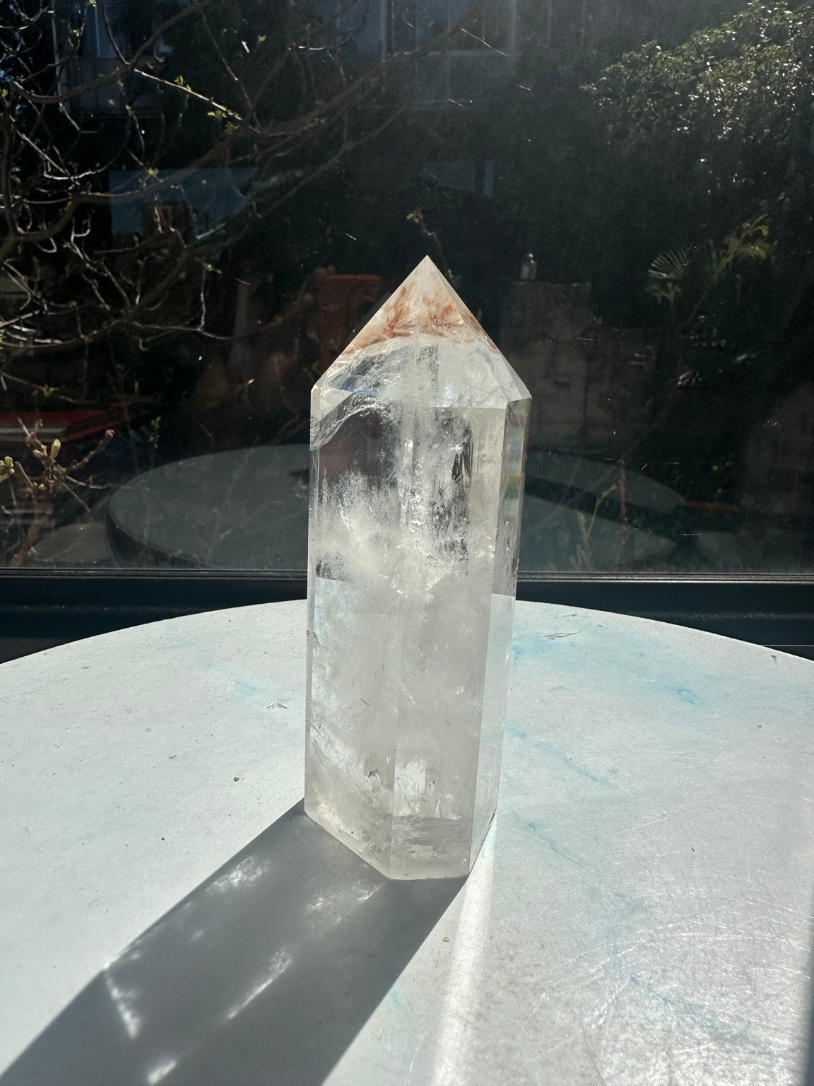 Brazilian clear quartz tower number 3, 467g and 13.2cm tall, standing on its flat base in sunlight. Clear upper facets show brown inclusions near the point and white veils through the body.