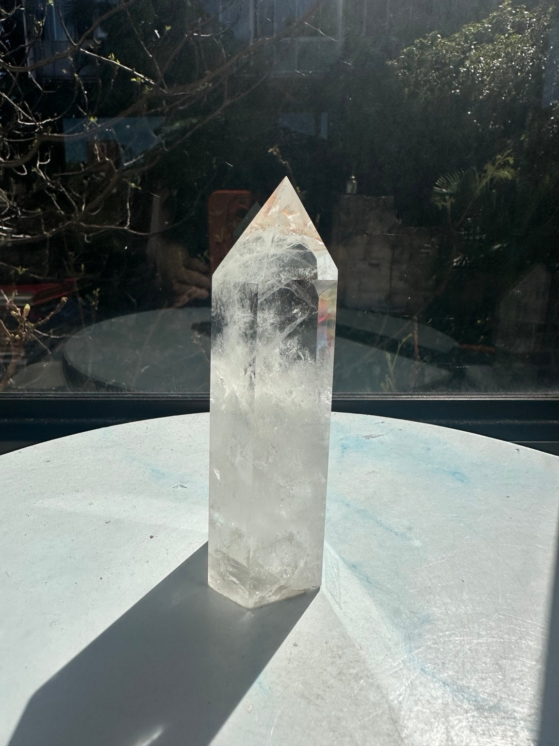 Side view of the exact 467g Brazilian clear quartz point, showing a subtle rainbow reflection along the right facet, internal fractures and a cloudy white lower section.