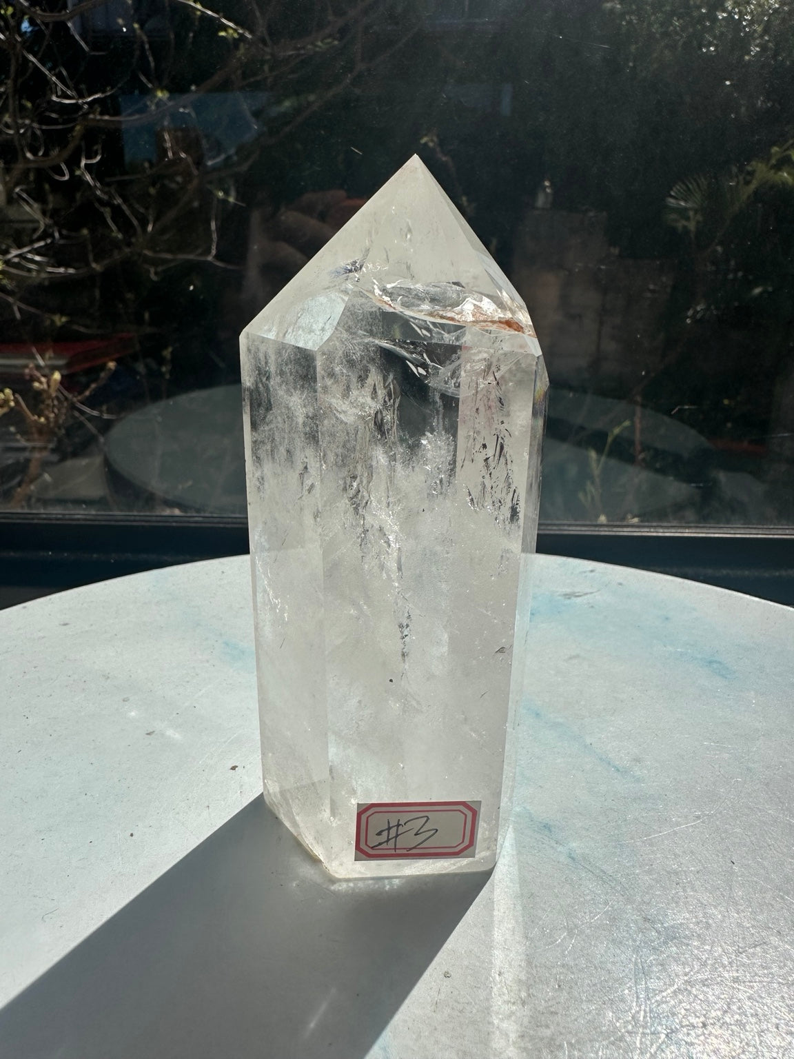 Front view of Brazilian clear quartz tower number 3 with its identification label at the base, transparent upper facets and white internal textures in bright sunlight.