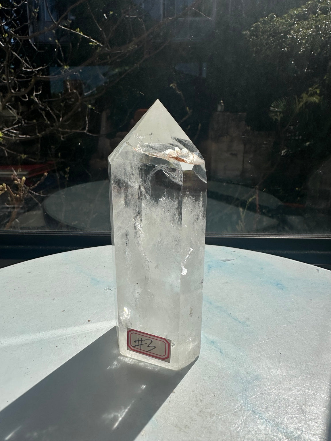 Angled front view of the 13.2cm Brazilian quartz tower with its number 3 label, polished edges, brown inclusion near the point and a long shadow on the display surface.