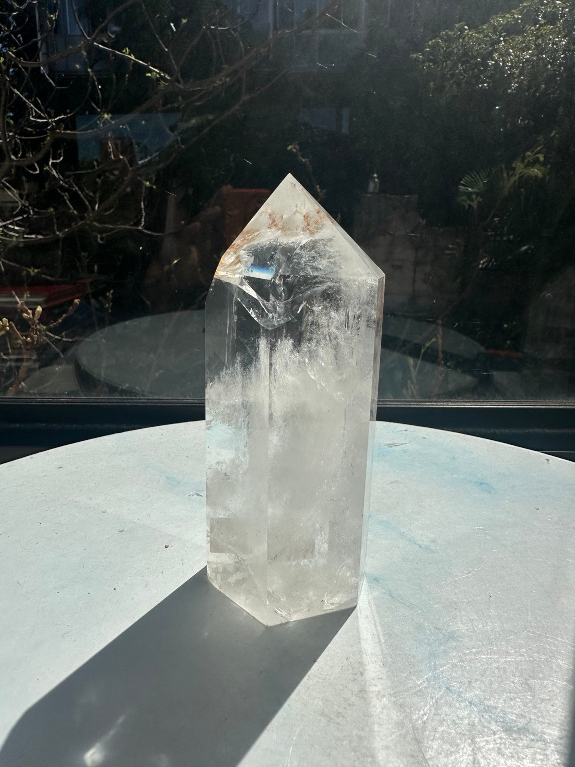 Rotated view of the Brazilian clear quartz tower with a small blue rainbow reflection near the upper facet, warm brown inclusions and white internal veils.
