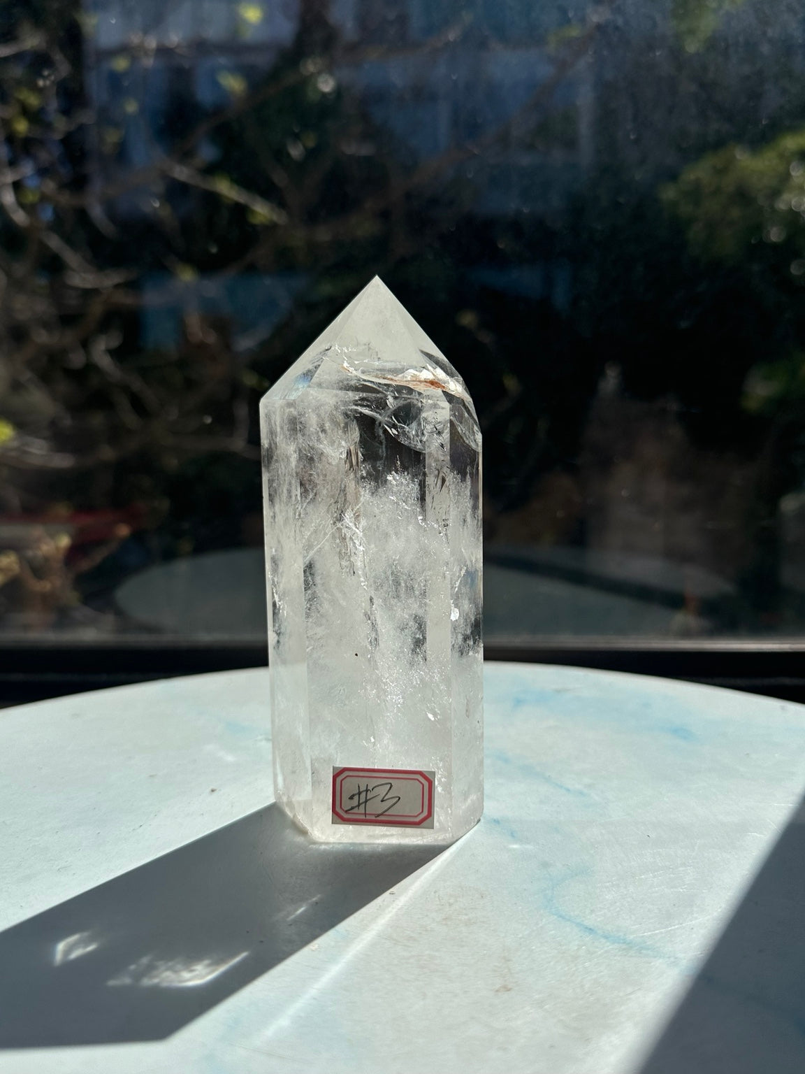Portrait of the 467g Brazilian clear quartz tower number 3 standing upright, with the complete tip and base visible against a softly blurred dark window background.