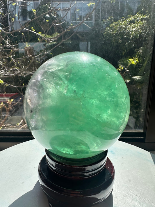 Large mint-green fluorite sphere from Madagascar on its included display stand. The 14cm, 3.9kg polished crystal ball shows translucent green areas, cloudy veils and internal lines in sunlight.