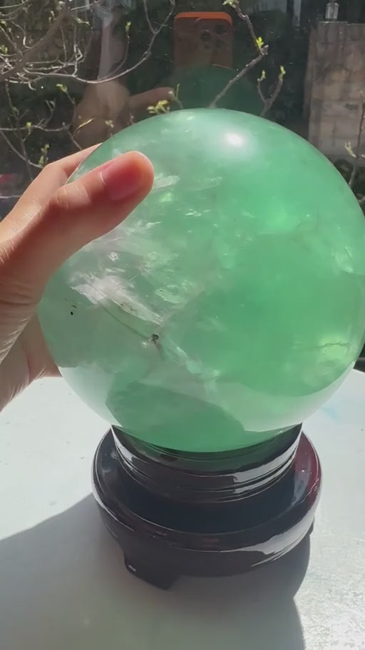 Exact 14cm, 3.9kg Madagascar green fluorite sphere rotating on its included display stand in natural sunlight, showing the translucency, veils and internal inclusions.