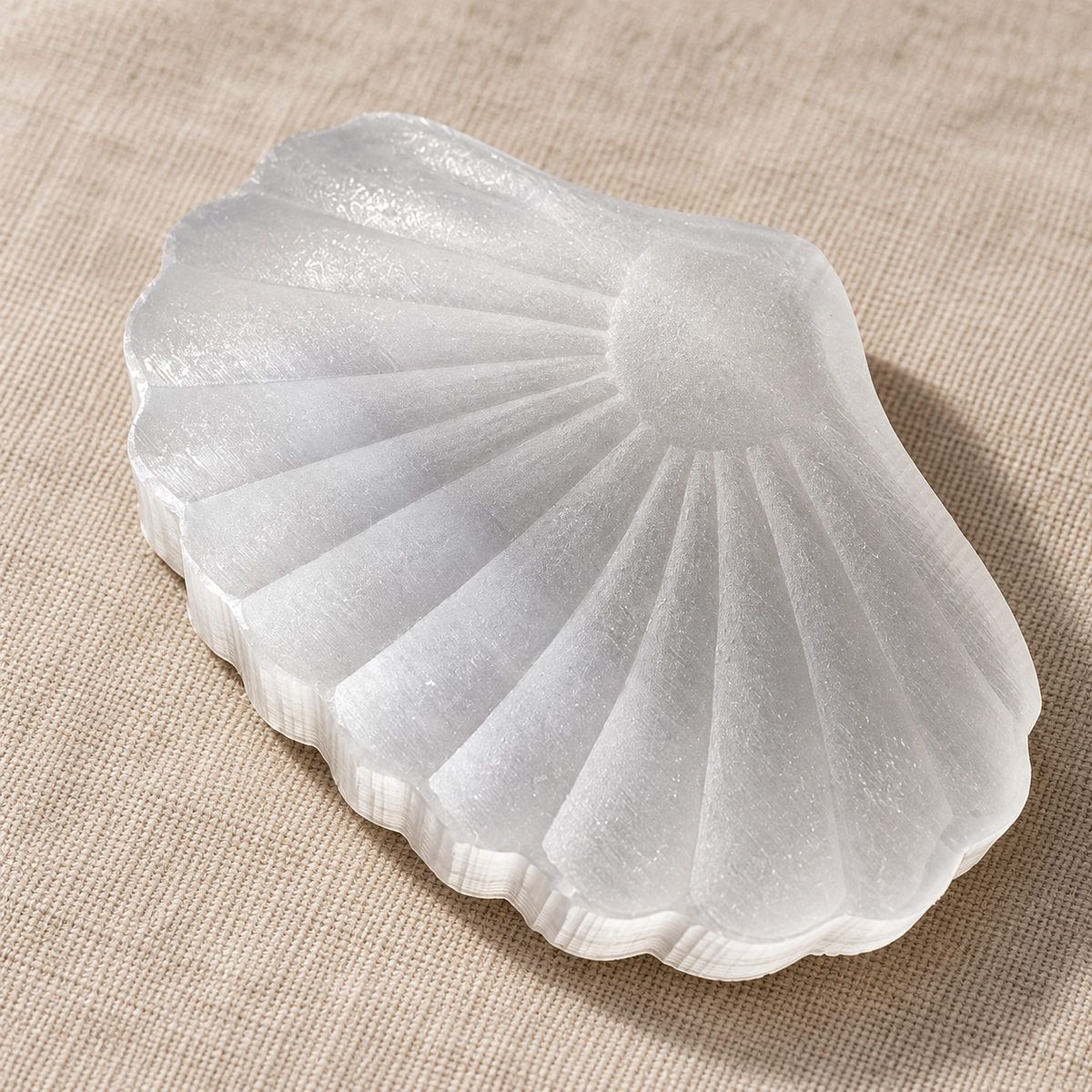 Angled close-up of a selenite shell carving on pale linen, showing the fan-shaped ridges and striped crystal edge.